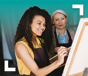 A smiling young woman paints on a canvas. There is an older instructor behind her watching her work.