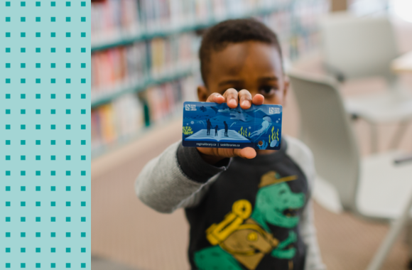 A little boy holds up a blue library card 