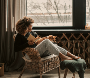 A woman reading on a wicker chair in front of a window. She has a dog in her lap and her feat are on a stool.