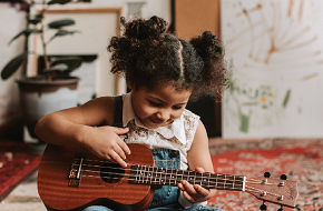 A little girl around age 5 plays a ukulele in her home.